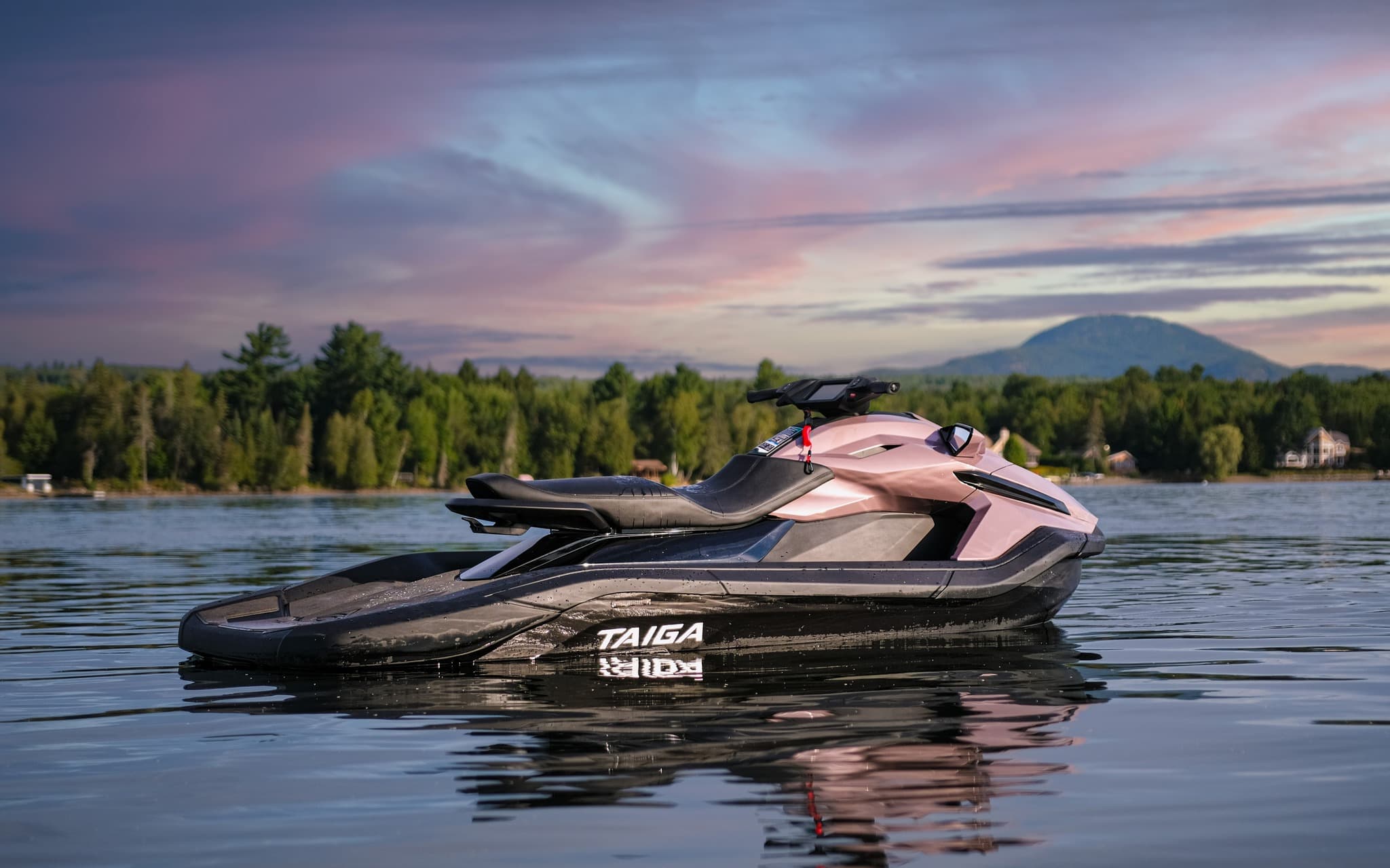 Taiga Orca WX3 electric personal watercraft floating on a calm lake, with a forested shoreline and mountain under a colorful sky.