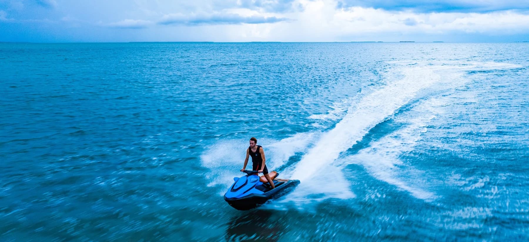 Rider operating a blue personal watercraft across open ocean, leaving a broad white wake under a partly cloudy sky.