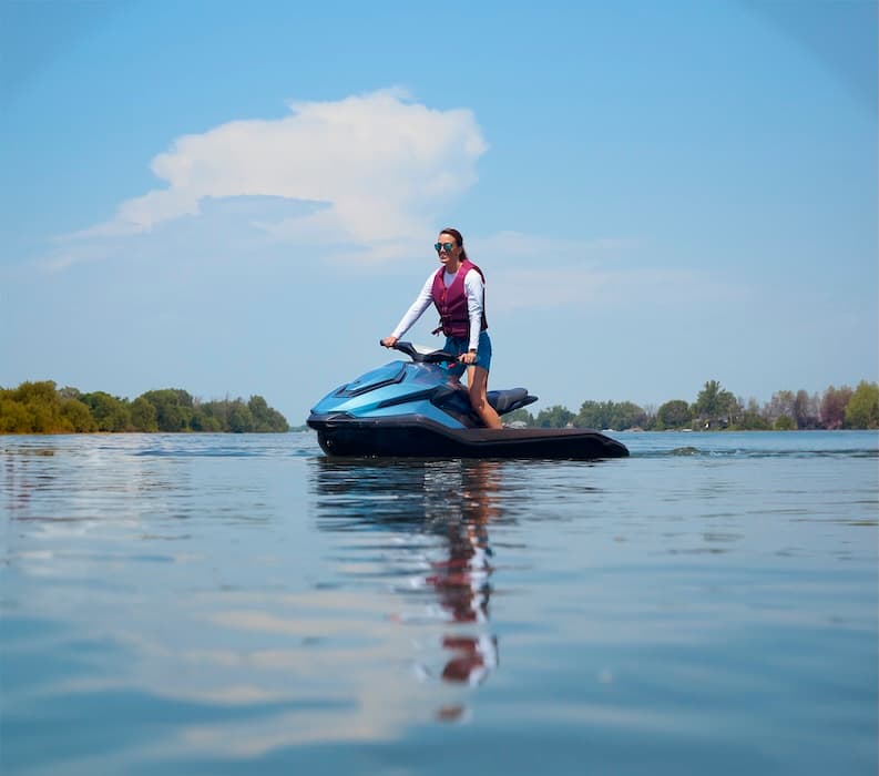 Rider operating a blue Taiga Orca WX3 personal watercraft across a calm lake with trees and a partly cloudy sky.