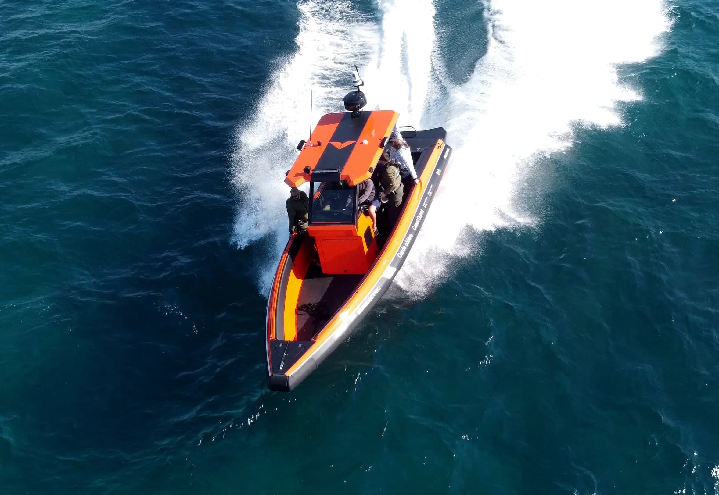 Orange-and-black patrol boat speeding across deep blue water, viewed from above, with two people aboard and a broad white wake.