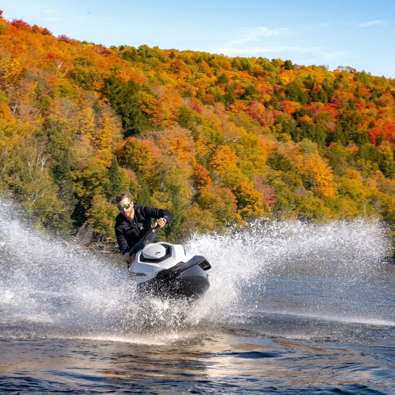 White Taiga Orca P2 electric personal watercraft turning on a lake, splashing beneath an autumn-colored forested hillside.
