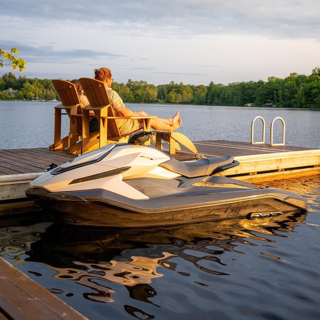 Taiga Orca P2 electric personal watercraft docked beside a lake, with two seated people and a swim ladder in the background.