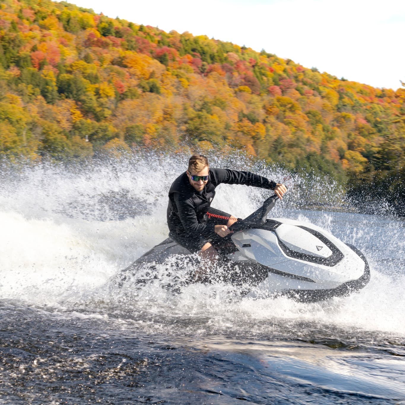 Rider turning on a white Taiga Orca P2 electric personal watercraft across a lake, spraying water against an autumn-colored hillside.