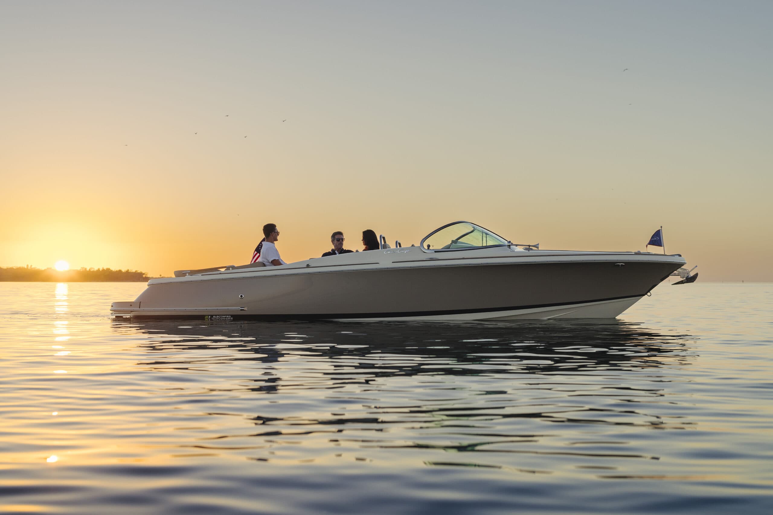 Chris Craft electric bowrider at low speeds on calm water at sunset, with several passengers aboard.