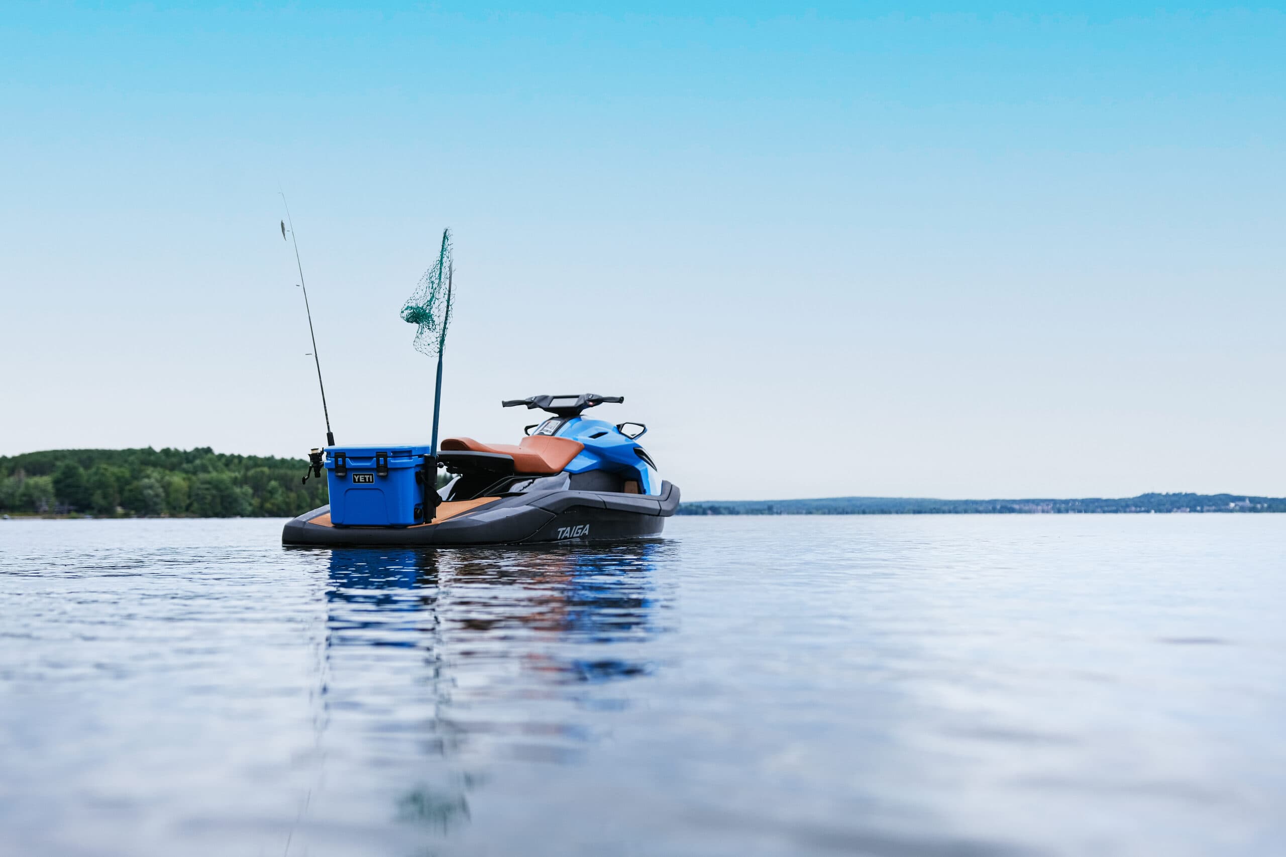 Taiga Orca personal watercraft floating on a calm lake with fishing gear, forested shoreline, and clear blue sky.