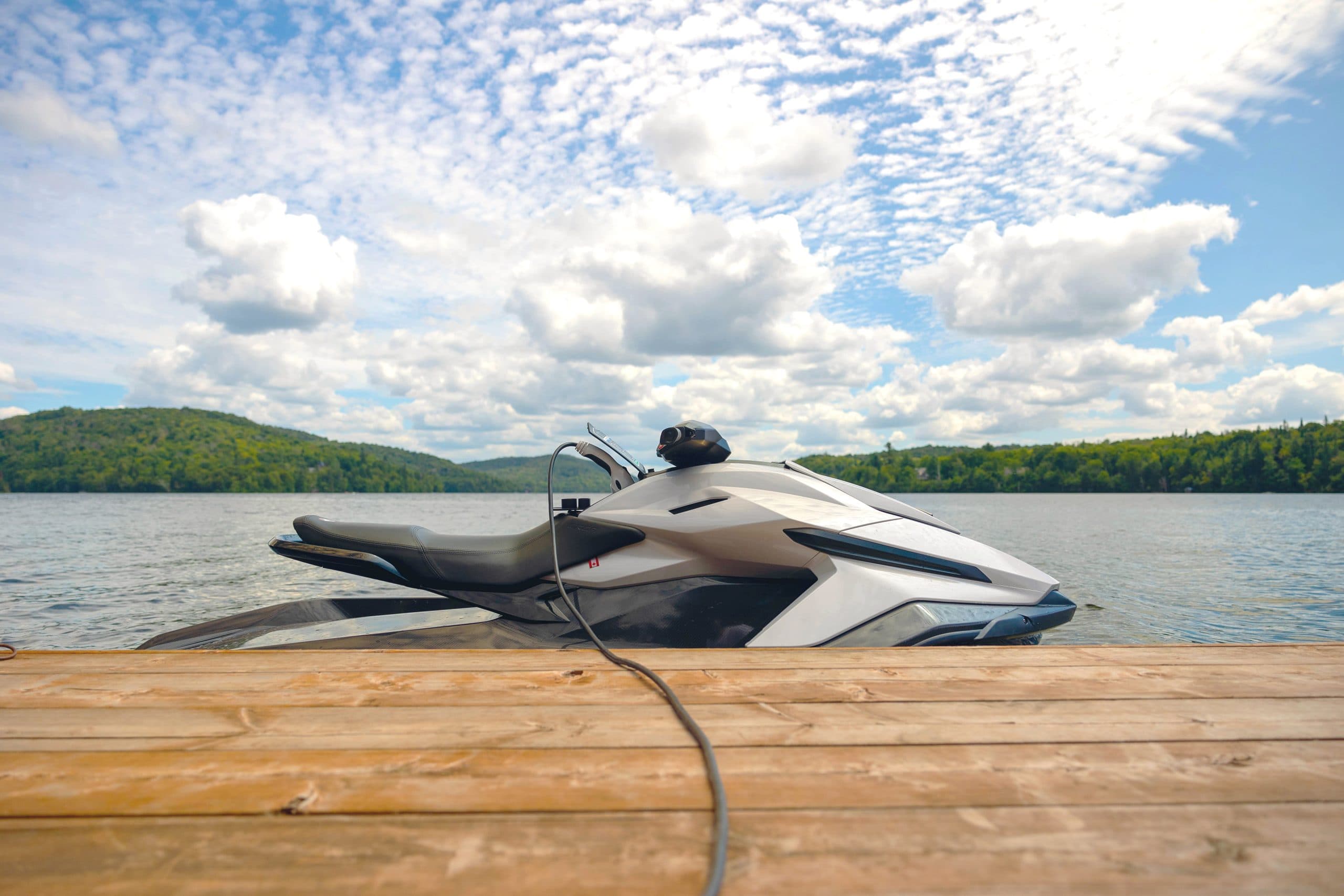 White Taiga Orca P2 electric personal watercraft docked and charging on a wooden pier beside a calm lake, with forested hills and clouds in the background.