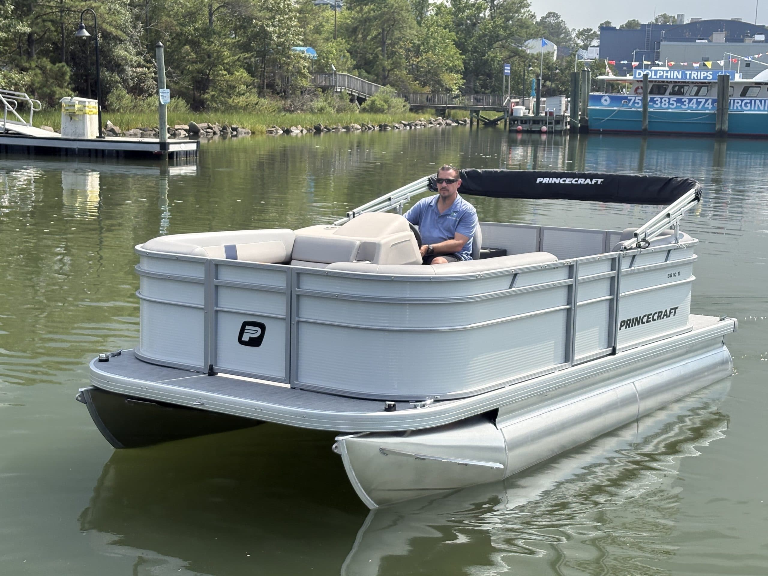 Princecraft Brio E17 white electric pontoon boat cruising on calm inland water, with a seated captain and docked boats in the background.