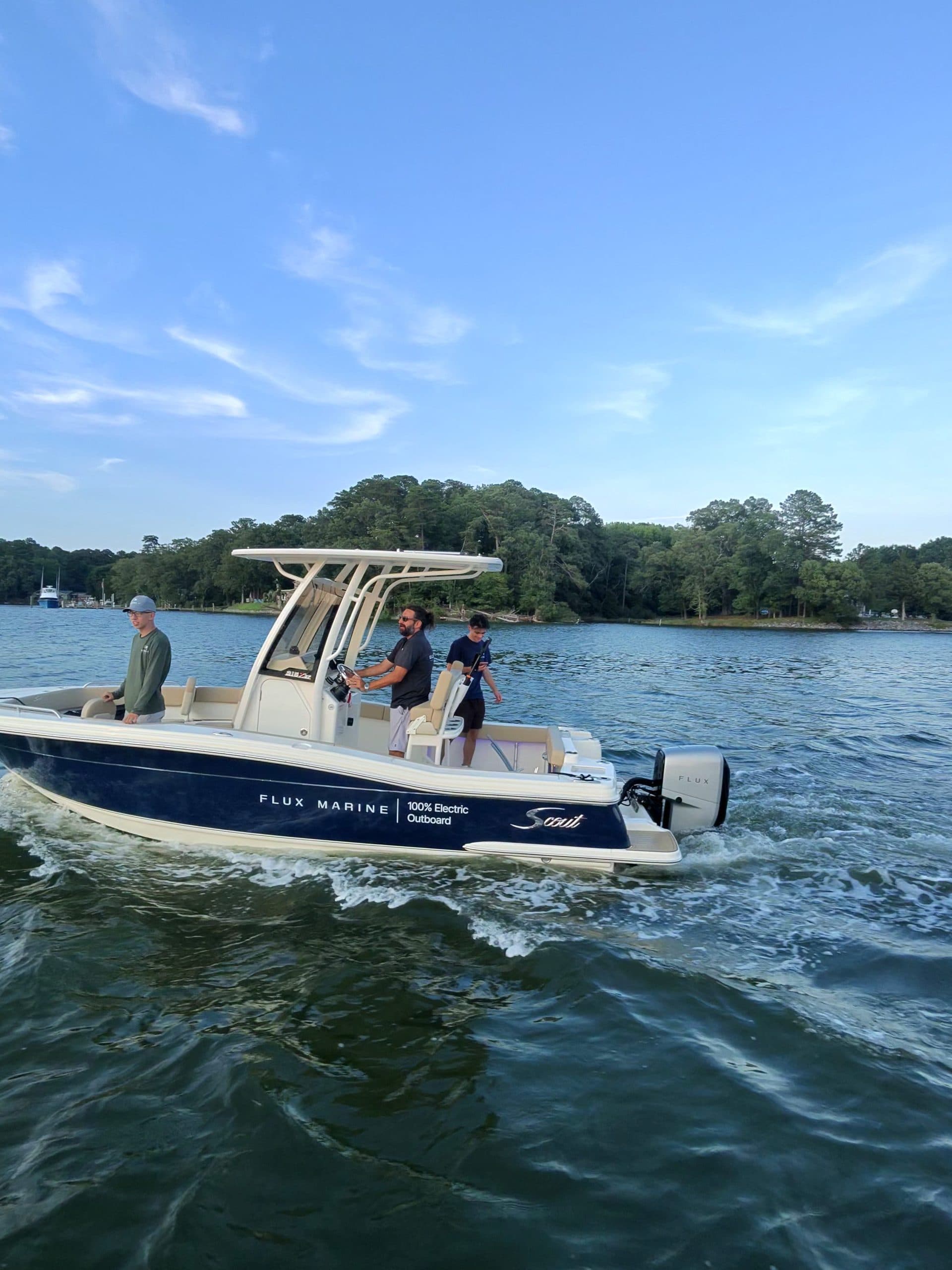 Scout 215 XSF center-console boat cruising on calm water, with three people aboard and a wooded shoreline in the background.