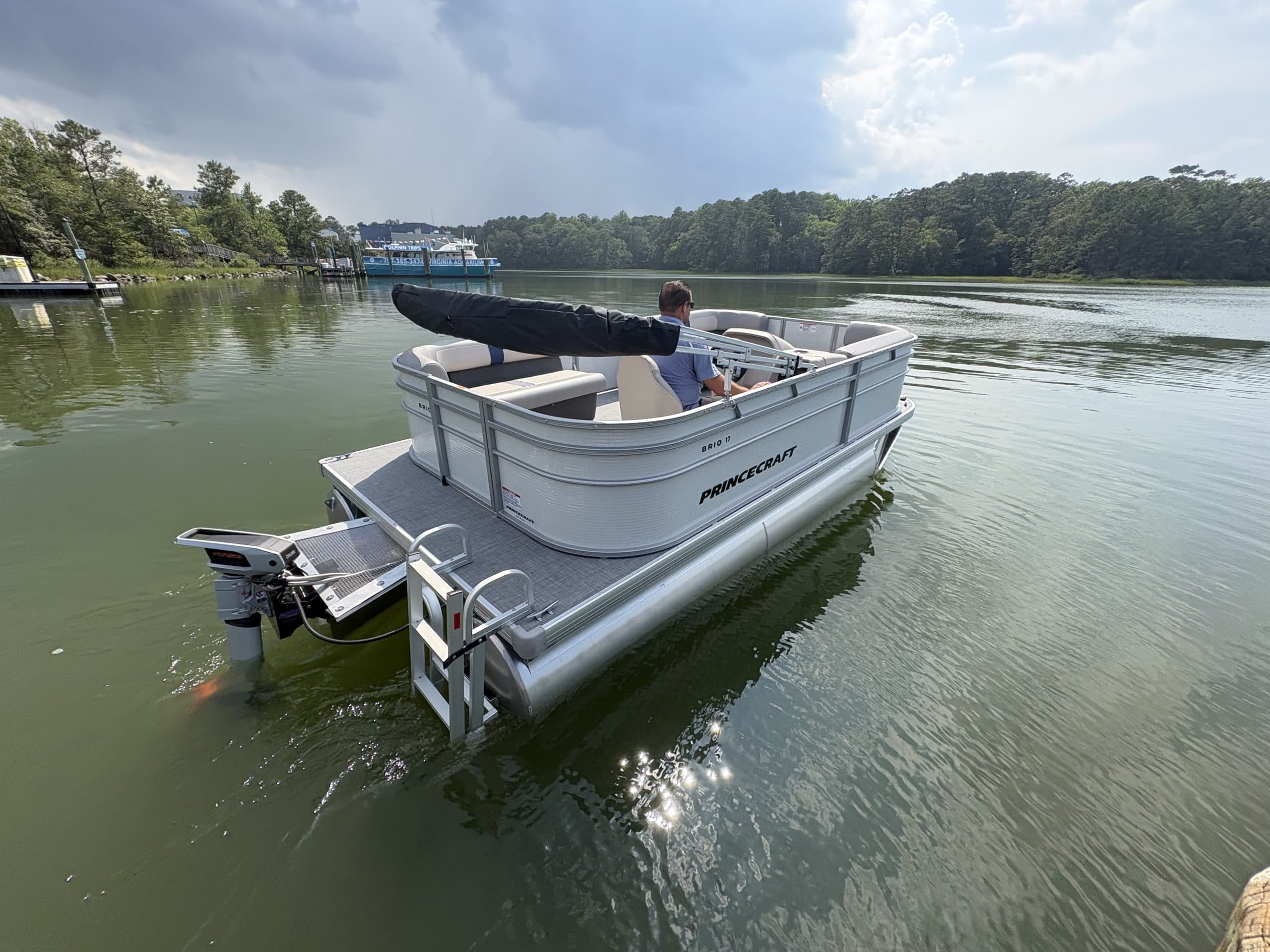 Princecraft Brio E17 electric pontoon boat on a calm lake, viewed from the rear with its outboard motor and boarding ladder visible.