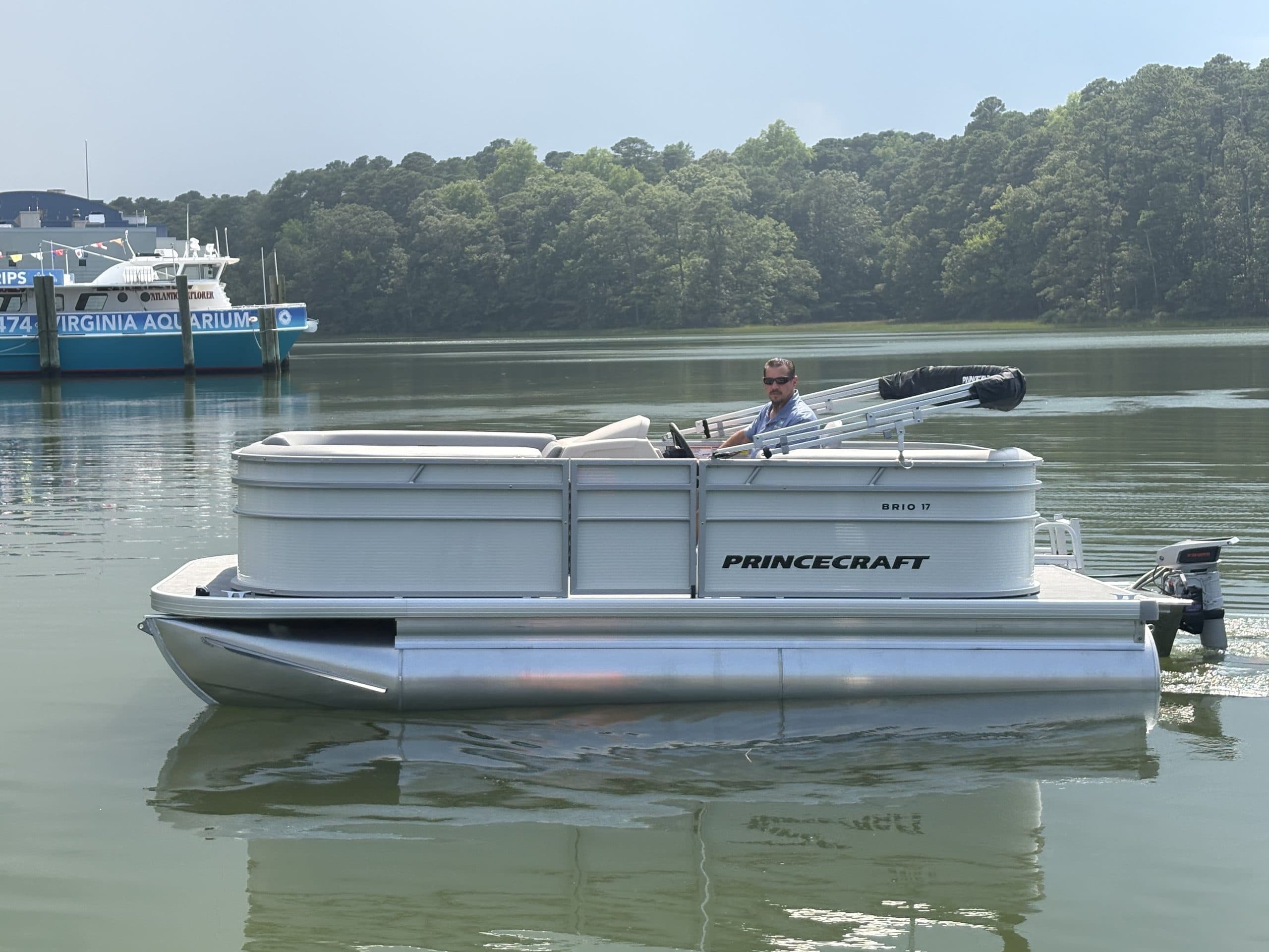 Princecraft Brio E17 pontoon boat with an outboard motor cruising on calm water near a tree-lined shore.