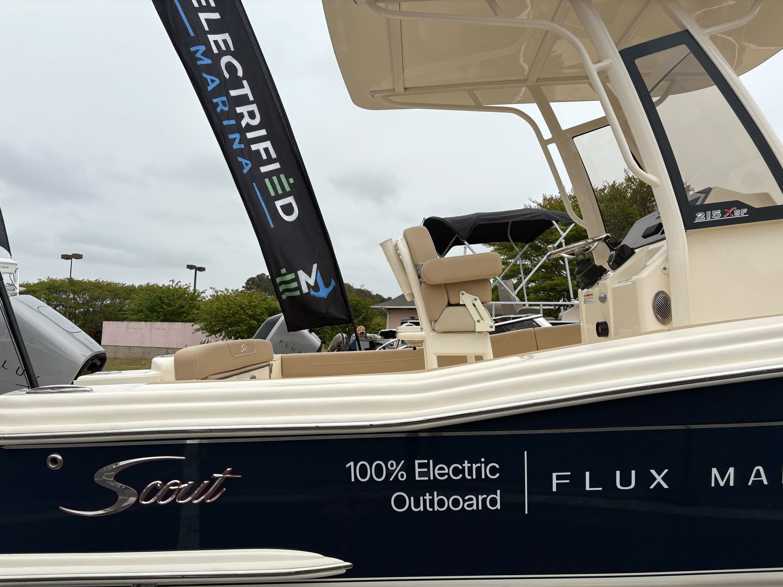 Close-up of a Scout 215 XSF electric center-console boat, showing the dark hull, “100% Electric Outboard” branding, seating, console, and T-top. Electrified Marina flag is seen in the background