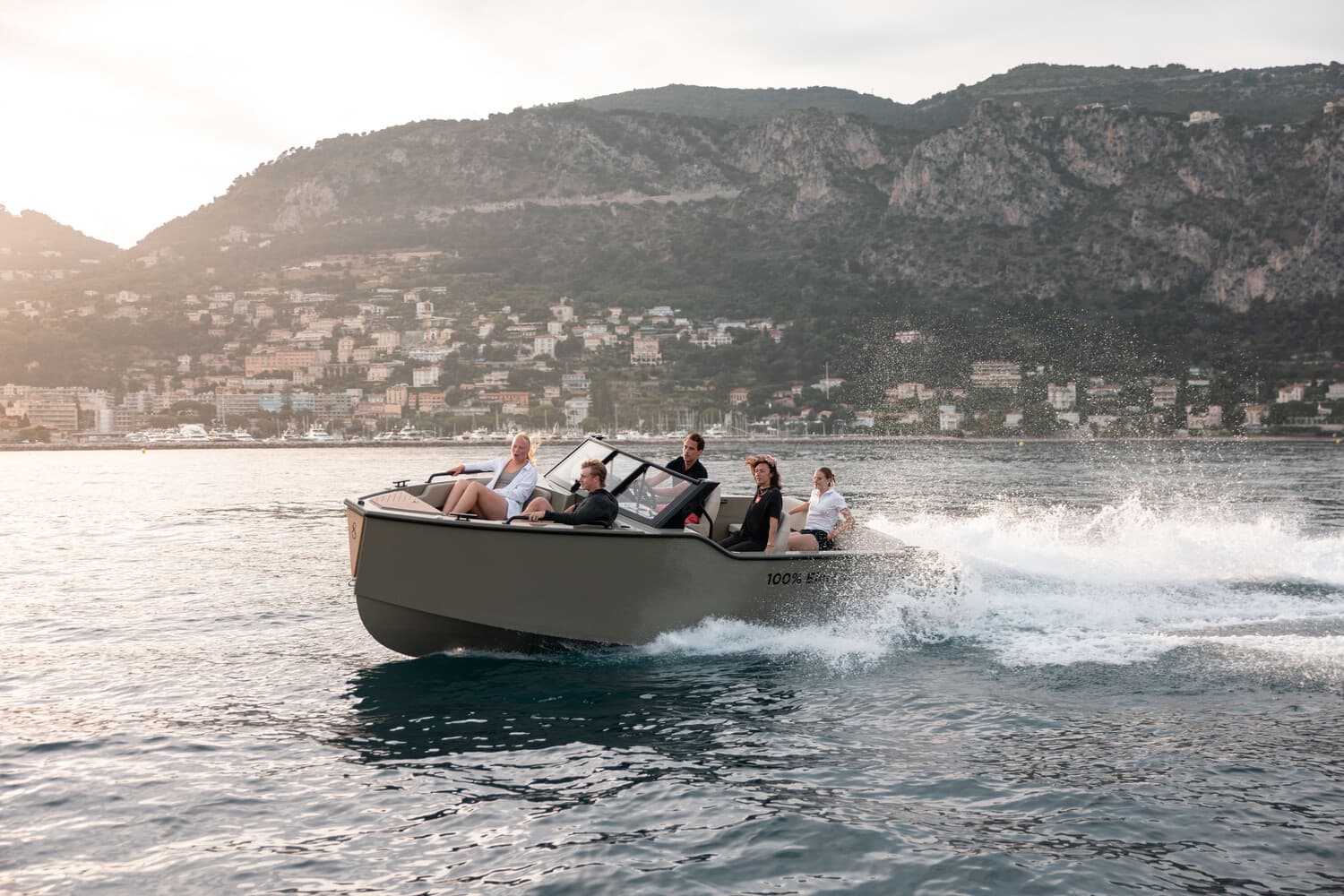 X Shore 1 electric boat carrying passengers across a lake, with a mountainous shoreline and coastal town in the background.