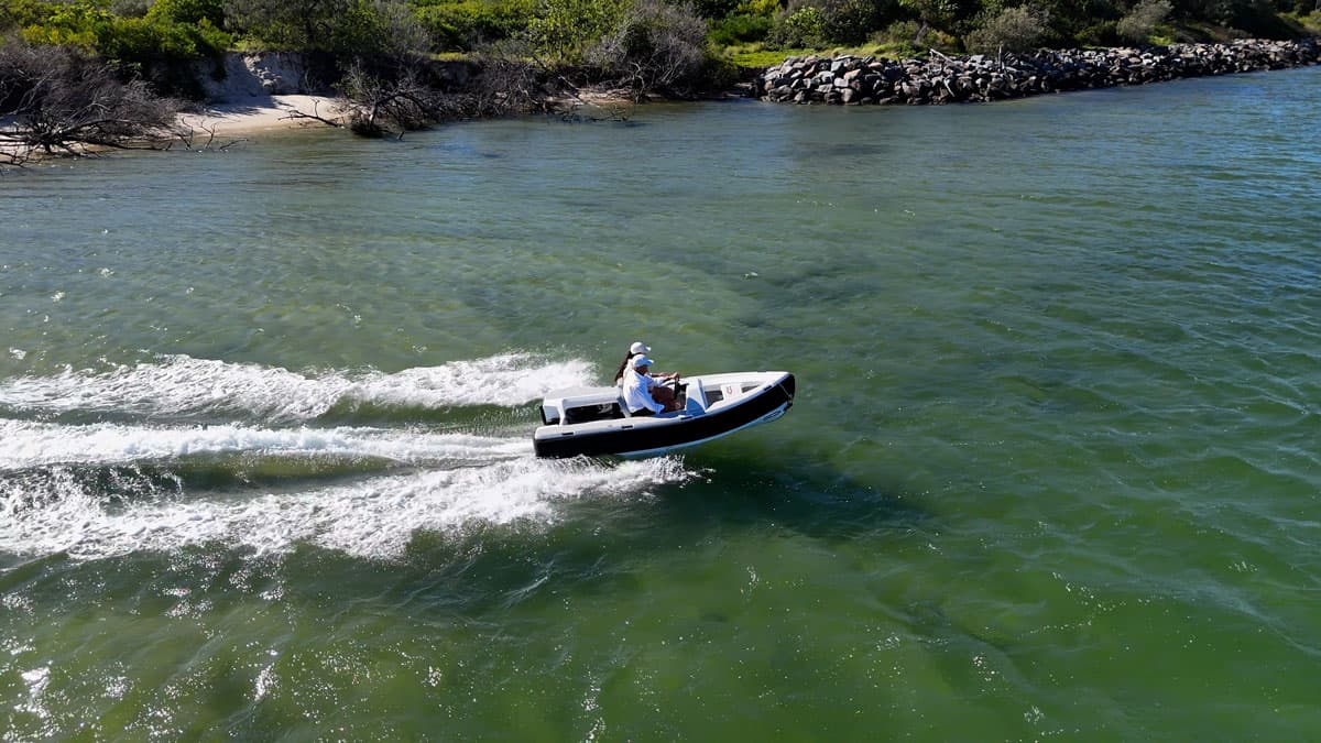 Two people in a white tender going fast speeds in the water with rocky coastline in the background